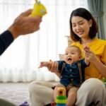 A woman is holding a baby and smiling. The baby is playing with a toy. The scene is warm and happy