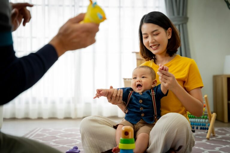A woman is holding a baby and smiling. The baby is playing with a toy. The scene is warm and happy